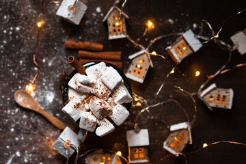 Winter hot drink: hot chocolate with marshmallows, cinnamon, anise. Christmas lights in the shape of houses. Decorative snow, dark wooden background. Festive mood, cozy atmosphere. Flat lay top view