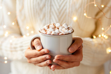 Young girl in knitted white wool sweater is holding a mug with hot chocolate or coffee with marshmallow. Christmas lights on, cozy holiday atmosphere, aromatherapy for cold winter season. Closeup