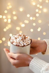 Young girl in knitted white wool sweater is holding a mug with hot chocolate or coffee with marshmallow. Christmas lights on, cozy holiday atmosphere, aromatherapy for cold winter season. Closeup