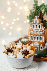 Delicious homemade baked gingerbread house with white icing. Rustic style, white wooden background, candles and christmas lights as decor. Cozy home atmosphere, festive mood, holiday inspiration