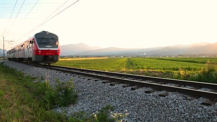Passenger train passing by on countryside railroad. Beautiful summer sunset with village in the distance. Railway between farming fields