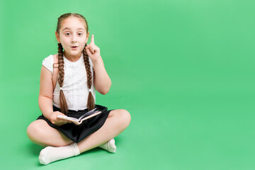 The little school girl on the floor with book shows her forefinger up on green background