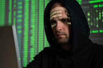 Male hacker working on a computer while green code characters reflect on his face in a dark office...