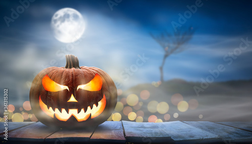 One spooky halloween pumpkin, Jack O Lantern, with an evil face and eyes on a wooden bench, table with a misty night full moon background with space for product placement.
