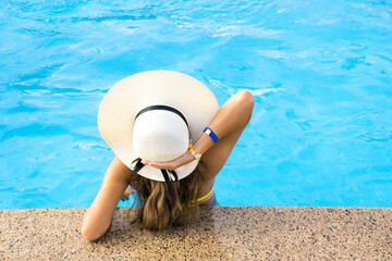 Fototapeta na wymiar Back view of young woman with long hair wearing yellow straw hat relaxing in warm summer swimming pool with blue water on a sunny day.