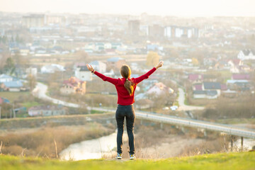 Fototapeta na wymiar Young woman standing outdoors raising her hands enjoying city view. Relaxing, freedom and wellness concept.