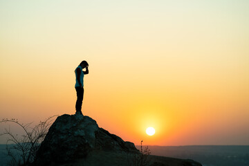 Silhouette of a woman hiker standing alone on big stone at sunset in mountains. Female tourist on high rock in evening nature. Tourism, traveling and healthy lifestyle concept.