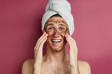Busy men prefer minimalist, easy and quick facial treatments. A  laughing man with a white terry towel on the head is applying brown scrubbing mask on his face and looking straight.