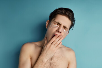 Busy men prefer minimalist, easy and quick facial treatments. A young man with pearl gel patches under his closed eyes is yawning and covering his mouth.