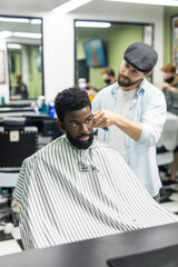 Happy young black man being trimmed with electric clipper machine in barbershop. Male beauty treatment concept. 