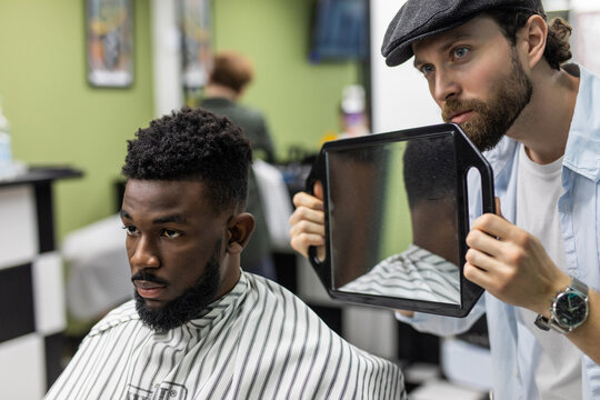 Portrait Of Handsome Black Man With Comb In Hair Looking In The Mirror At His New Haircut. Barber Hairdresser Showing Client His Work. Male Beauty Treatment Concept. Pretty Young African Guy Face