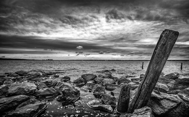 A black and white landscape scenery of rocks and wooden posts at a lakeside with dramatic clouds