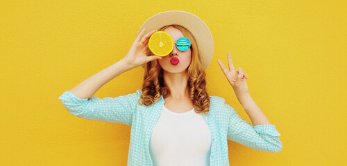 Summer portrait of young woman covering her eyes with fruits slice of orange blowing red lips sending sweet air kiss wearing straw hat on yellow background