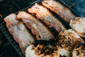 Slices of thin bacon on a grill. Cooking pork on coals. Toasting burger buns. Outdoor Food. Picnic.