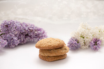 Three oatmeal cookies. In the background, lilac and white lilac flowers. White background
