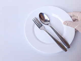 A hand with white latex glove holds a metal spoon, a fork, and an empty plate. Isolated on a white background.