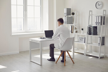 Doctor online. Male doctor works talking video call with patient using bast shoe while sitting at desk in back view clinic office.