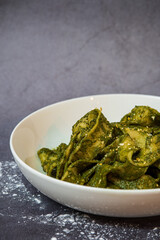 Fresh homemade gluten-free pappardelle pasta with pesto sauce in a white bowl on dark table surface with flour on the table,  very broad and flat pasta noodles. with copy space