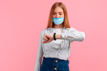 young smiling woman in a medical mask, looking at the clock on her hand, on a pink background