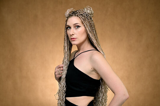 Photo of a caucasian fashionable woman in black blouse with long afro dreadlocks hair in studio. Pretty model posing on a beige background standing right in front of the camera.