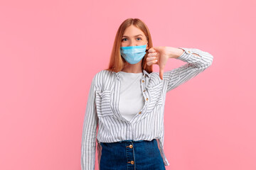 disgruntled annoyed unhappy young woman in a medical protective mask, the woman gives a thumbs down as an unfriendly gesture isolated on a pink background