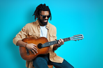 Charming young African man in casual wear playing the guitar and smiling while sitting against blue background