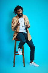 Full length of trendy young African man in casual wear looking at camera and pointing you while sitting on stool against blue background