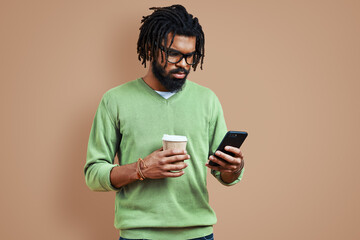 Busy young African man in smart casual clothing using smart phone while standing against brown background