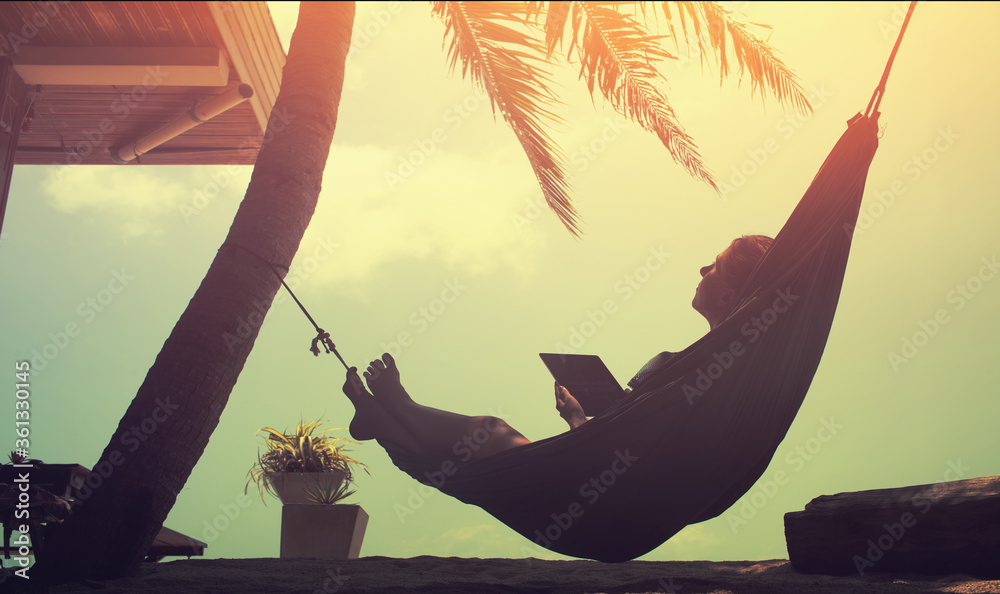 Wall mural woman an using a digital tablet while relaxing in a hammock on a sand tropical summer beach