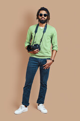 Full length of young African photographer in casual clothing looking at camera while standing against brown background