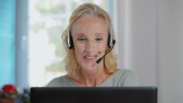 Happy mature telemarketer woman with laptop attending call with headset