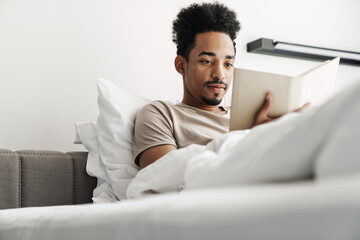 Photo of serious african american man reading book while lying in bed