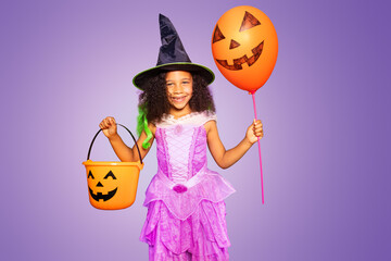 Cute little girl in Halloween dress hold orange balloon and candy bucket over purple background