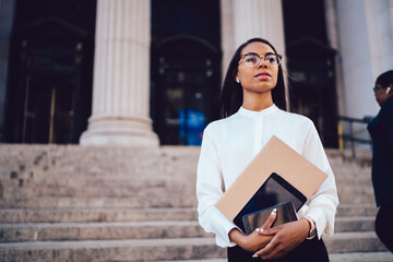 Confident dark skinned student of faculty of law looking staight while holding modern smartphone and touch pad in hands strolling near university building.Serious female office worker with folder