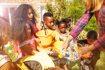 Close-up view of many children in spooky costumes receive Halloween candies from woman standing near house together outside