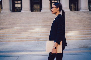 Side view of stylish African American businessman with folder in hands walking outdoors in city downtown, Female entrepreneur in elegant wear moving on street. Copy space for financial information