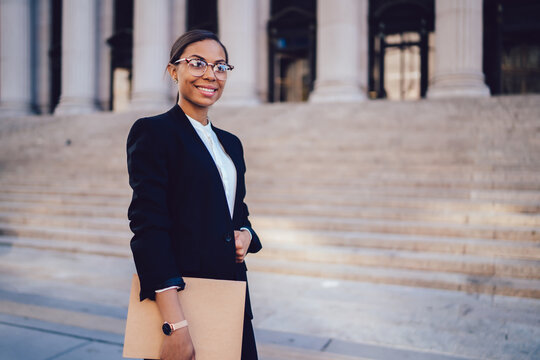 Portrait Of African American Businesswoman With Successful Career Dressed In Formal Wear Standing Against Architecture Office Building With Folder In Hands. Experienced Woman Entrepreneur Smiling