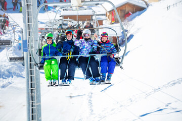 Group of skiers children sit on chairlift and wave hands going on the mountain to ski, sitting together smiling