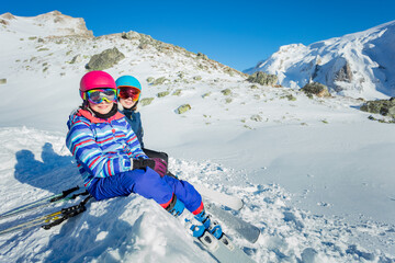 Fototapeta na wymiar Two happy girls sit on the snow in ski masks and helmets looking at the mountains