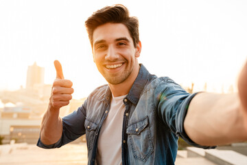 Photo of joyful young man showing thumb up while taking selfie