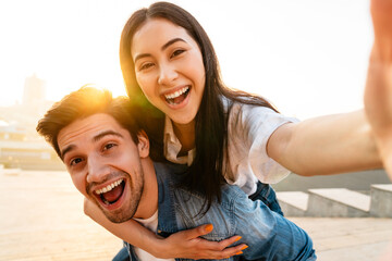 Image of multicultural couple piggybacking ride and taking selfie