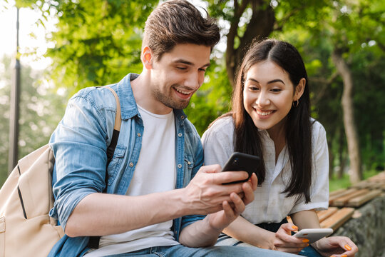 Image of pleased couple using mobile phones while sitting on bench