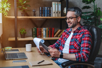 Obraz premium Middle aged caucasian executive reading documents and working on laptop while sitting at his desk in home office