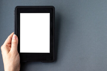 A modern black electronic book with a white blank empty screen in female hand on a gray paper background. Mockup tablet closeup with copy space