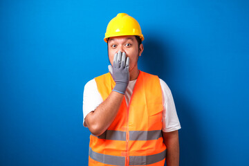 Asian worker wearing a helmet looks shocked hearing the news while closing his mouth with his hands against blue background