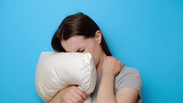 Tired sad young woman embraces pillow feels pain in neck after night sleep, awaken having painful sudden ache or stiffness, incorrect posture during sleep, isolated over blue studio background