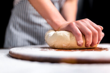 Woman kneading dough on wooden board sprinkled with flour with a black background. Concept of making dough for pizza. Close-up lock of a piece of dough being kneaded by a chef.