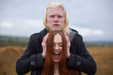 A handsome male albino model with an emotionless face in a black jacket holds his hands on the sides of a red-haired girl who screams against a wheat field. Unusual couple.