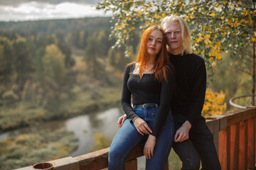 Happy couple guy albino and girl with bright red hair in dark clothes, sit on the fence leaning against each other against the background of the forest in the park