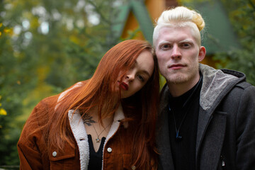 Portrait of an unusual couple. An albino man in a dark jacket and a girl with bright red hair in a corduroy jacket look to the camera against a backdrop of green trees
